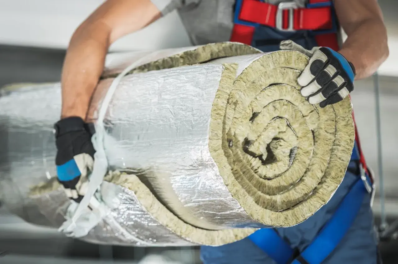 Budowlaniec w rękawicach niesie zrolowaną wełnę mineralną, owiniętą folią aluminiową.