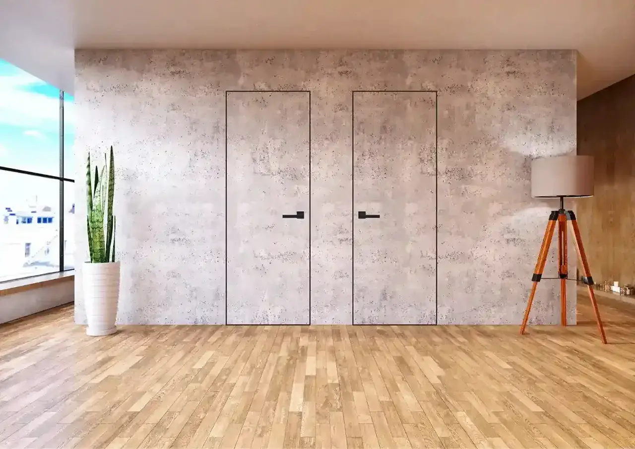 Dwa minimalistyczne, **drzwi ukryte w ścianie** z betonu architektonicznego, z czarnymi uchwytami. Obok stoi lampa i roślina.