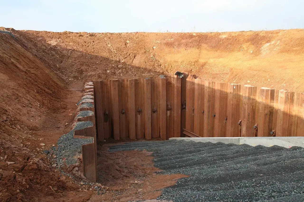 Konstrukcja oporowa typu larsen stabilizuje wykop. Widoczne są stalowe ścianki szczelne i nasypy.