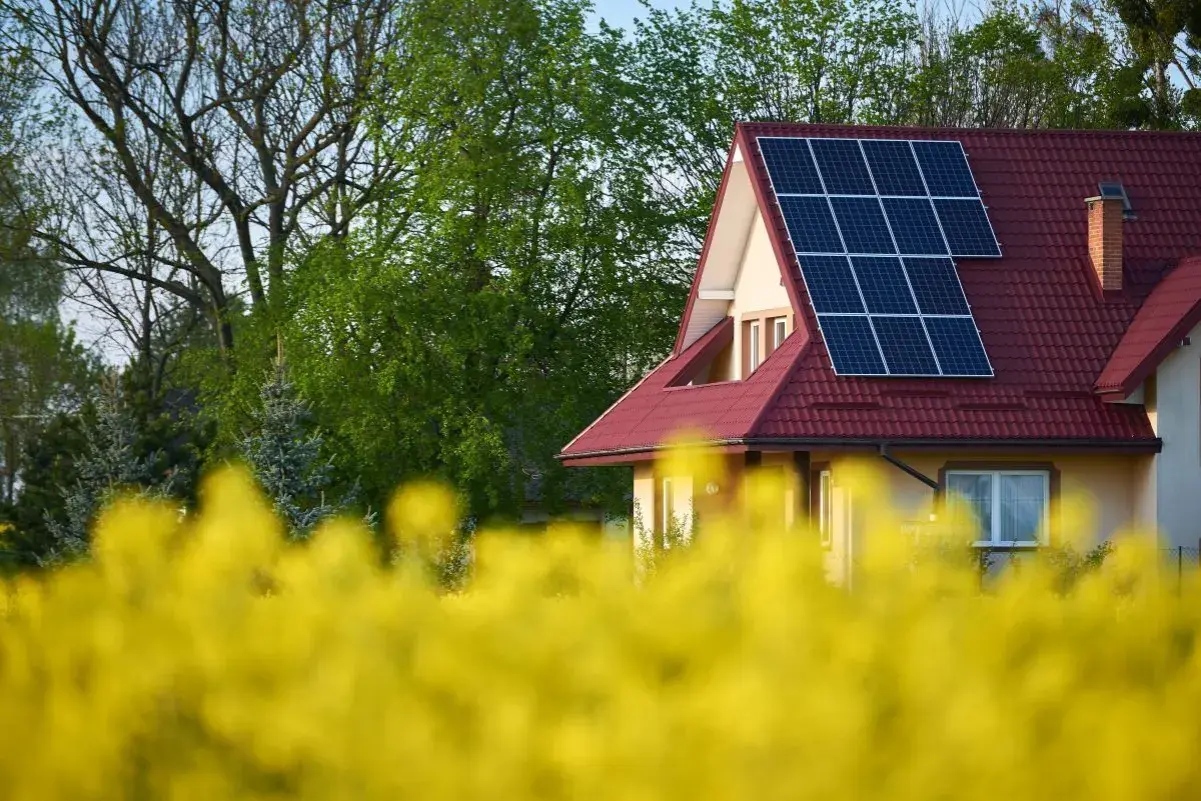 Dom z instalacją PV na czerwonym dachu, otoczony kwitnącym rzepakiem i zielenią drzew.