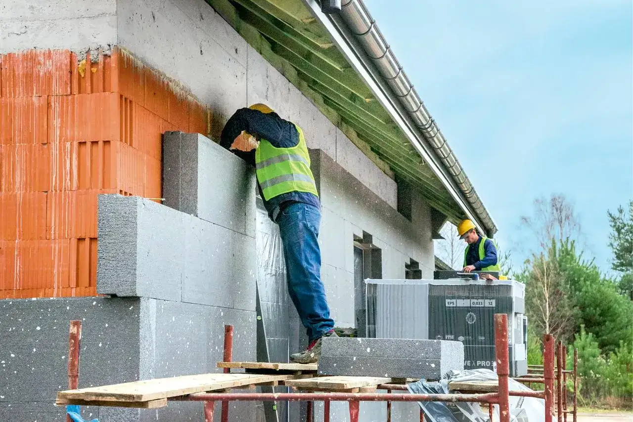 Budowlańcy montują płyty izolacyjne. Widać różne grubości styropianu na ścianie domu w trakcie ocieplania.