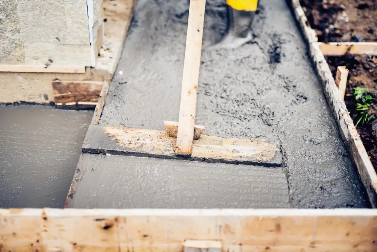 Prace budowlane w toku: świeży beton wylewany jest do szalunku, a pracownik w żółtych kaloszach wyrównuje go za pomocą drewnianej łaty. Projekt b20 nabiera kształtów.