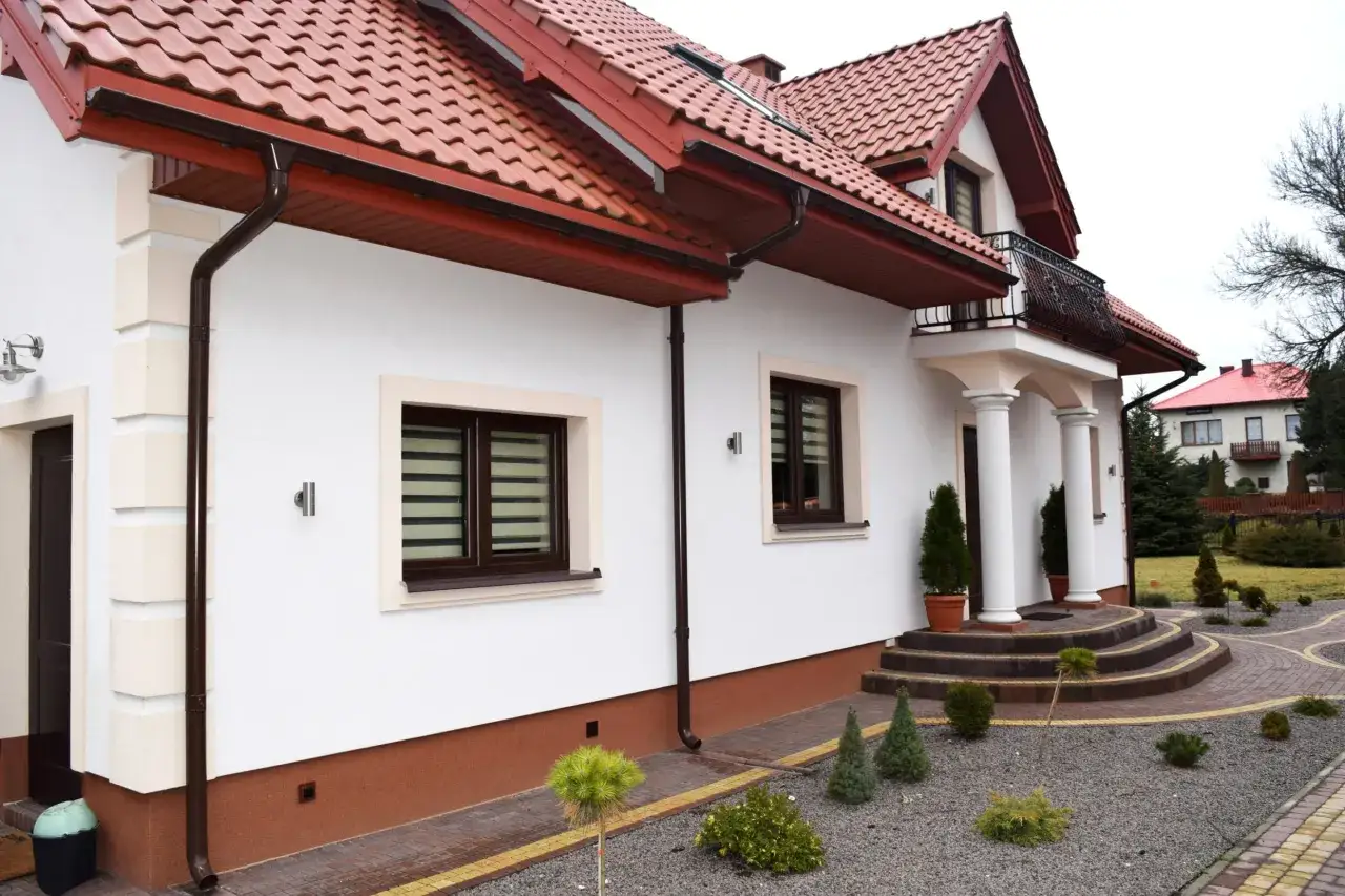 Nowoczesny dom z białymi ścianami i brązowym cokołem, ozdobiony kamiennymi elementami i zielenią.