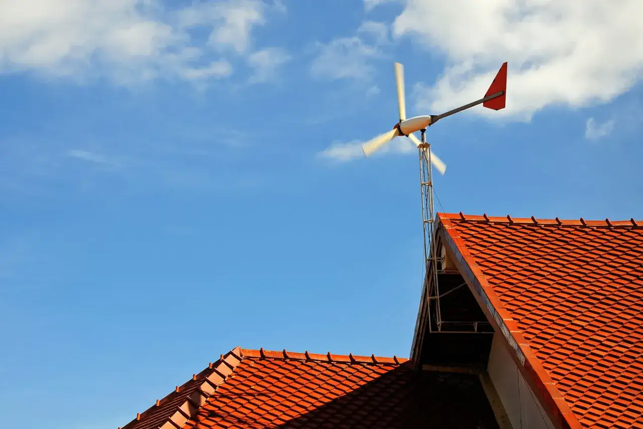 Mała elektrownia wiatrowa na dachu domu, z czerwonymi dachówkami i błękitnym niebem.