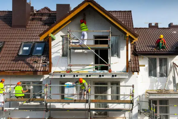 Pracownicy na rusztowaniach prowadzą termomodernizację domu, docieplając ściany i dach.