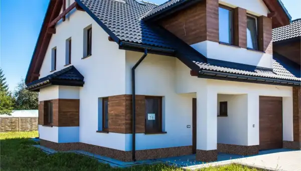 Nowoczesny dom z białym tynkiem, drewnianymi akcentami i ciemnym dachem. Uwagę przyciąga cokół z płytek imitujących kamień.