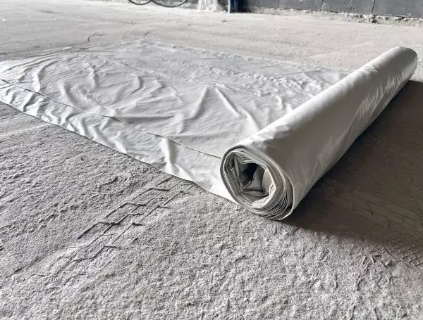 Biała folia budowlana rozwinięta na szarej, pylistej podłodze. Rolka folii leży po prawej stronie, tworząc cień.