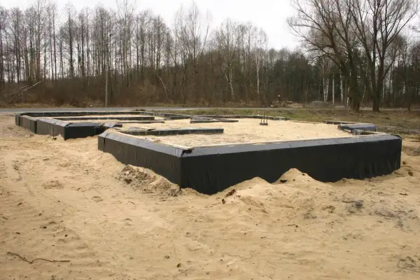Fundament domu w piasku, z czarnym lepikiem izolującym ściany fundamentowe. W tle las.