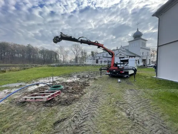 Prace przy studni głębinowej: dźwig z ramieniem pracuje nad wykończeniem, w tle budynek.