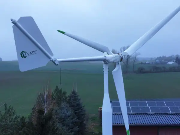 Biała elektrownia wiatrowa Falcon z zielonymi akcentami na tle zielonych pól i nieba. Obok widoczne panele słoneczne na dachu.