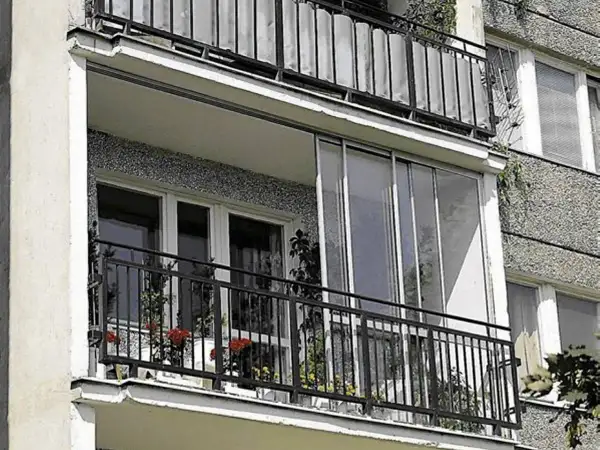 Zabudowa balkonu w bloku z przeszklonymi drzwiami i metalową balustradą. Na balkonie stoją doniczki z kwiatami.