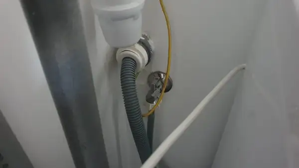 Szara rura odpływowa pralki podłączona do syfonu pod zlewem. Widoczny żółty przewód i biały kabel.