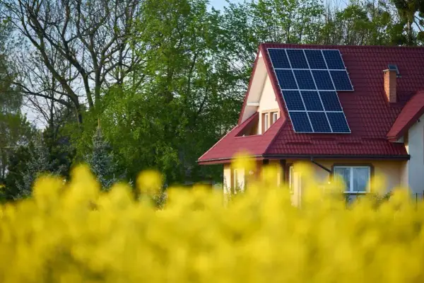 Dom z instalacją PV na czerwonym dachu, otoczony kwitnącym rzepakiem i zielenią drzew.