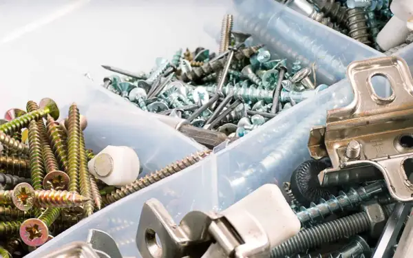 Pudełko z różnymi śrubkami, wkrętami i elementami metalowymi. Widać złote i srebrne śrubki, gwoździe, a także metalowe zawiasy.