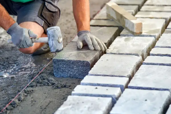 Brukarz układa kamienne płyty, używając młotka. Widać mokry, świeży beton, a obok suche płyty, sugerujące odpowiednie suchy beton proporcje.